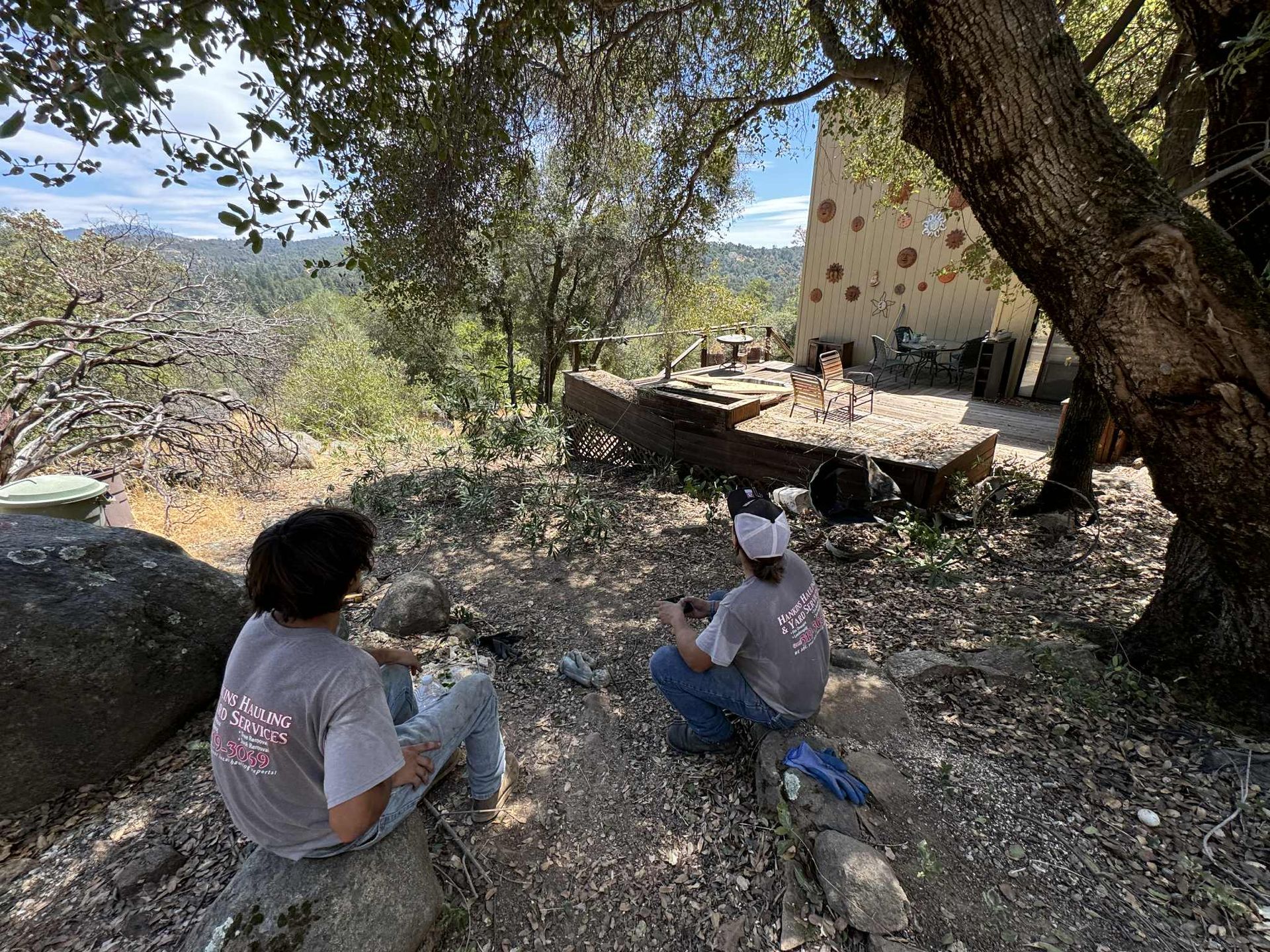 Two people sit on a hillside, looking towards a building. Trees and a view of mountains are in the background.