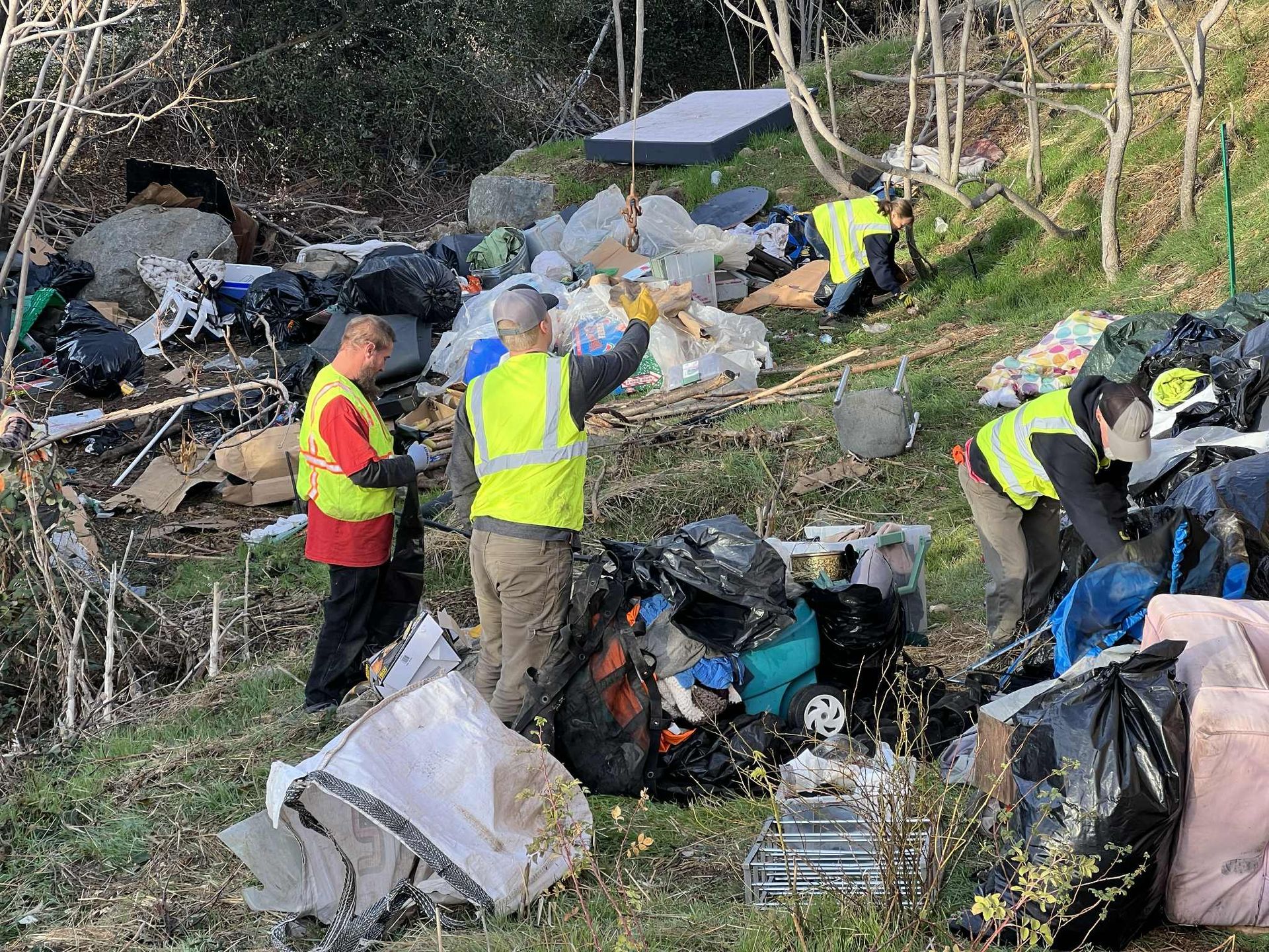Volunteers in reflective vests cleaning trash on a hillside, collecting garbage in black bags.