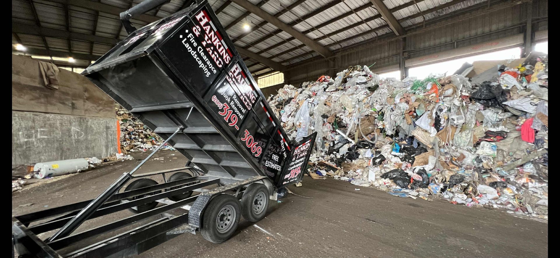 A black dump trailer unloading trash into a large pile inside a warehouse.