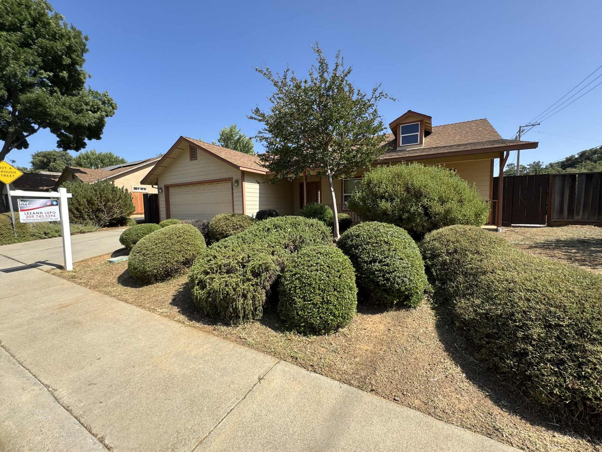 House with beige facade and brown roof, surrounded by green shrubs on a sunny day. A for sale sign is in the yard.