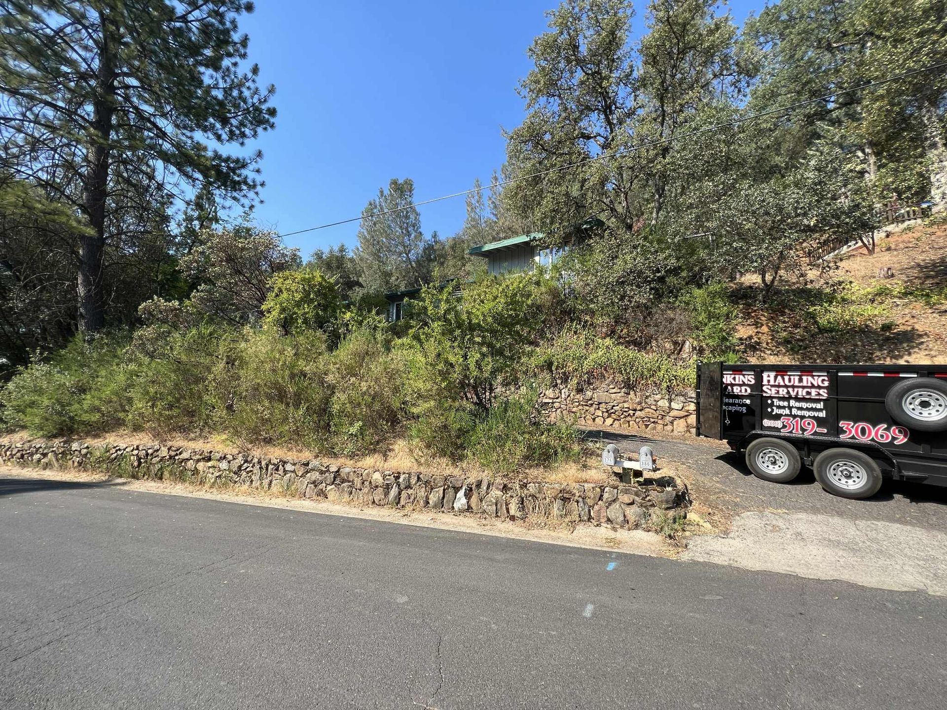 Roadside view of a green house surrounded by trees and bushes on a hill. A trailer is parked on the right.