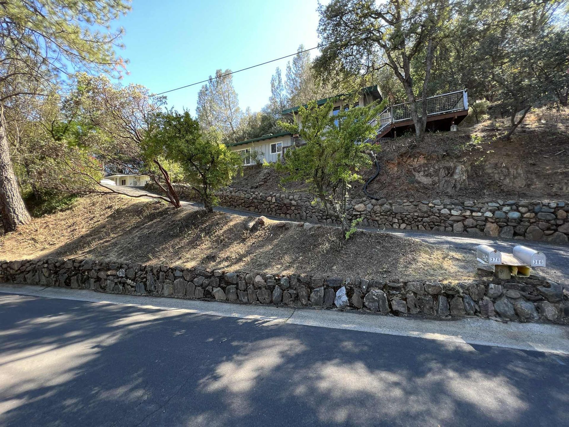 A hillside home with stone retaining walls, trees, and string lights on a sunny day.