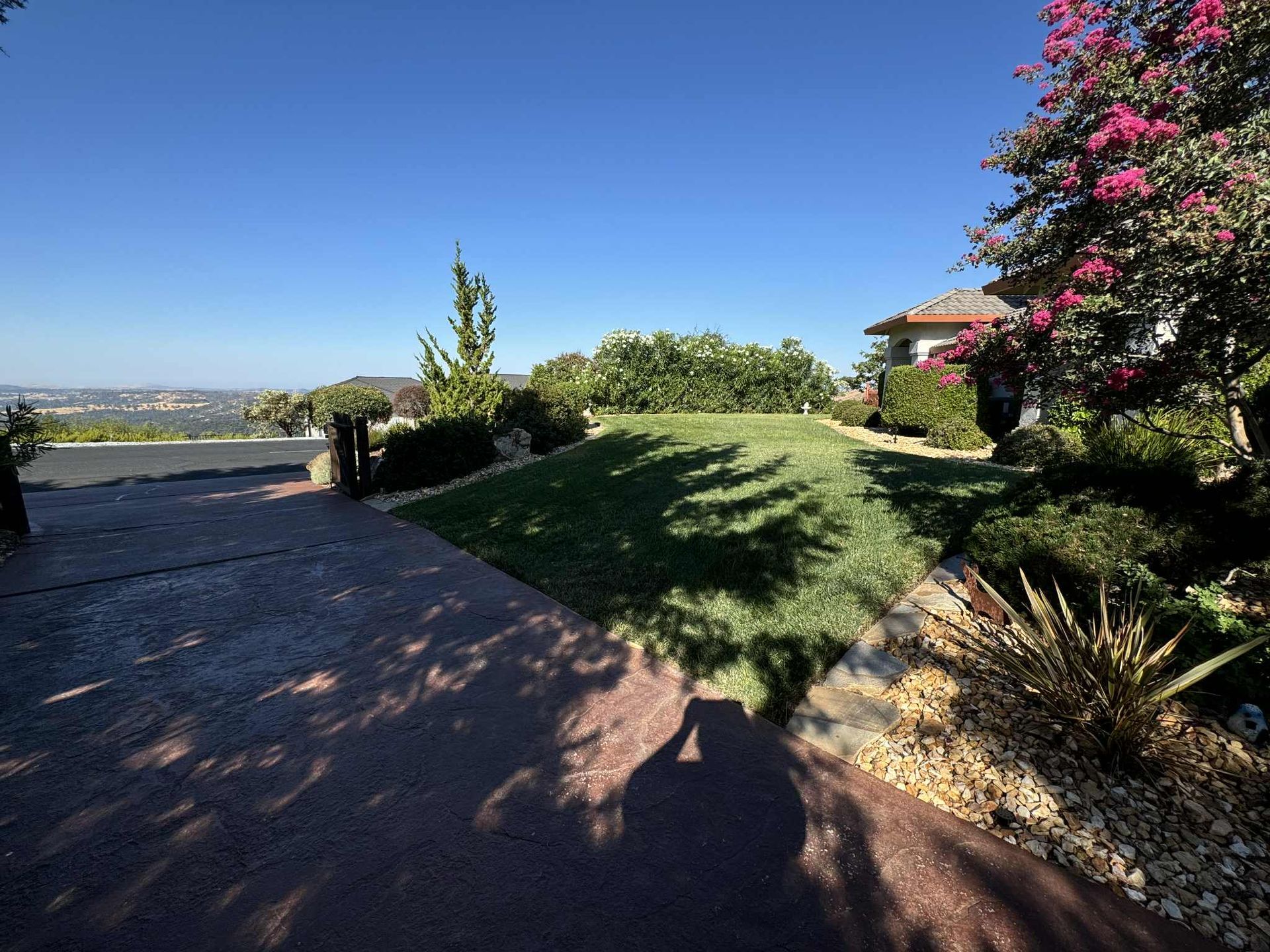 Driveway leads to a green lawn with trees and a house, overlooking a distant cityscape on a sunny day.