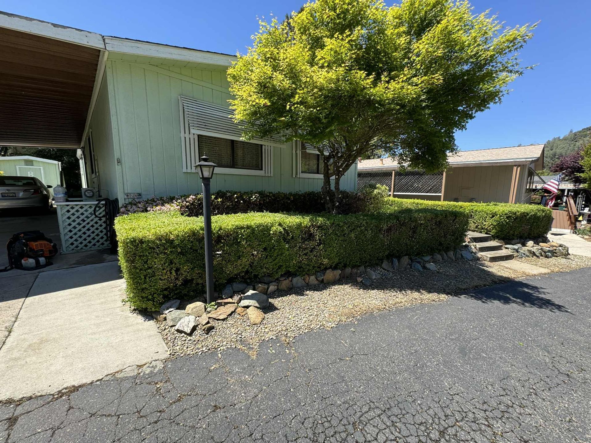 Light green house with manicured hedges, a tree, and a lamp post in front. Asphalt and gravel.