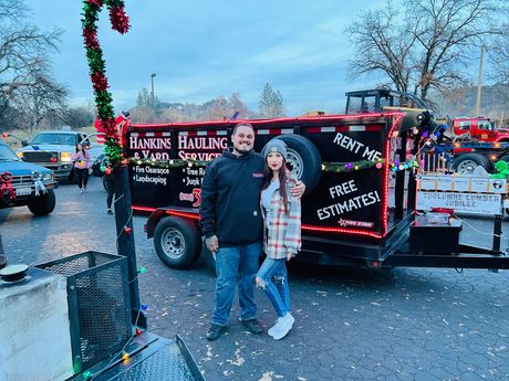 Couple standing in front of a decorated hauling trailer at a parade; dusk setting.
