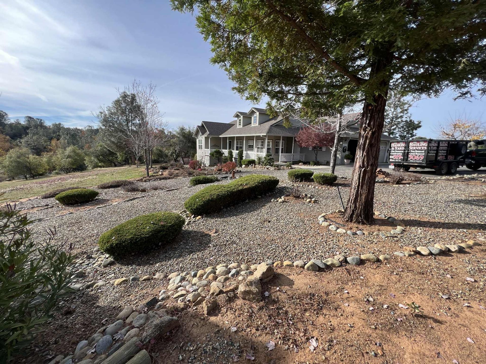 A house on a hillside with rock landscaping, bushes, and a tree. A truck is parked to the side.