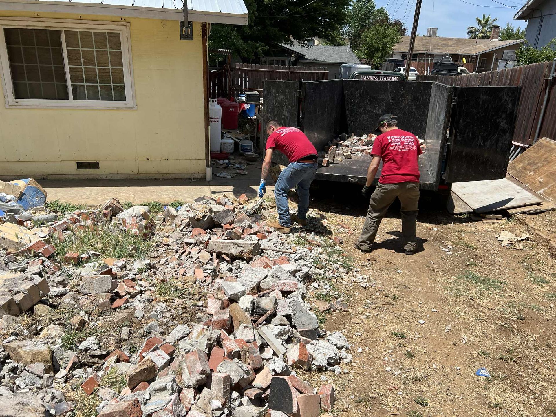 Two men in red shirts load debris into a dumpster near a house.