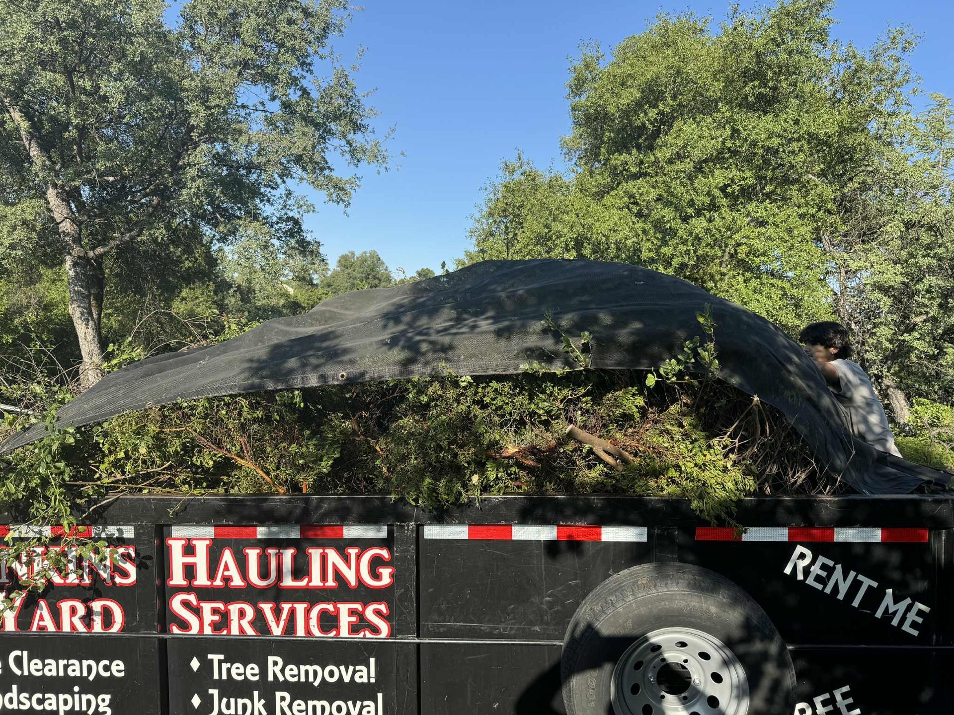 A trailer filled with yard waste, covered with a tarp. A person stands nearby, under a blue sky.