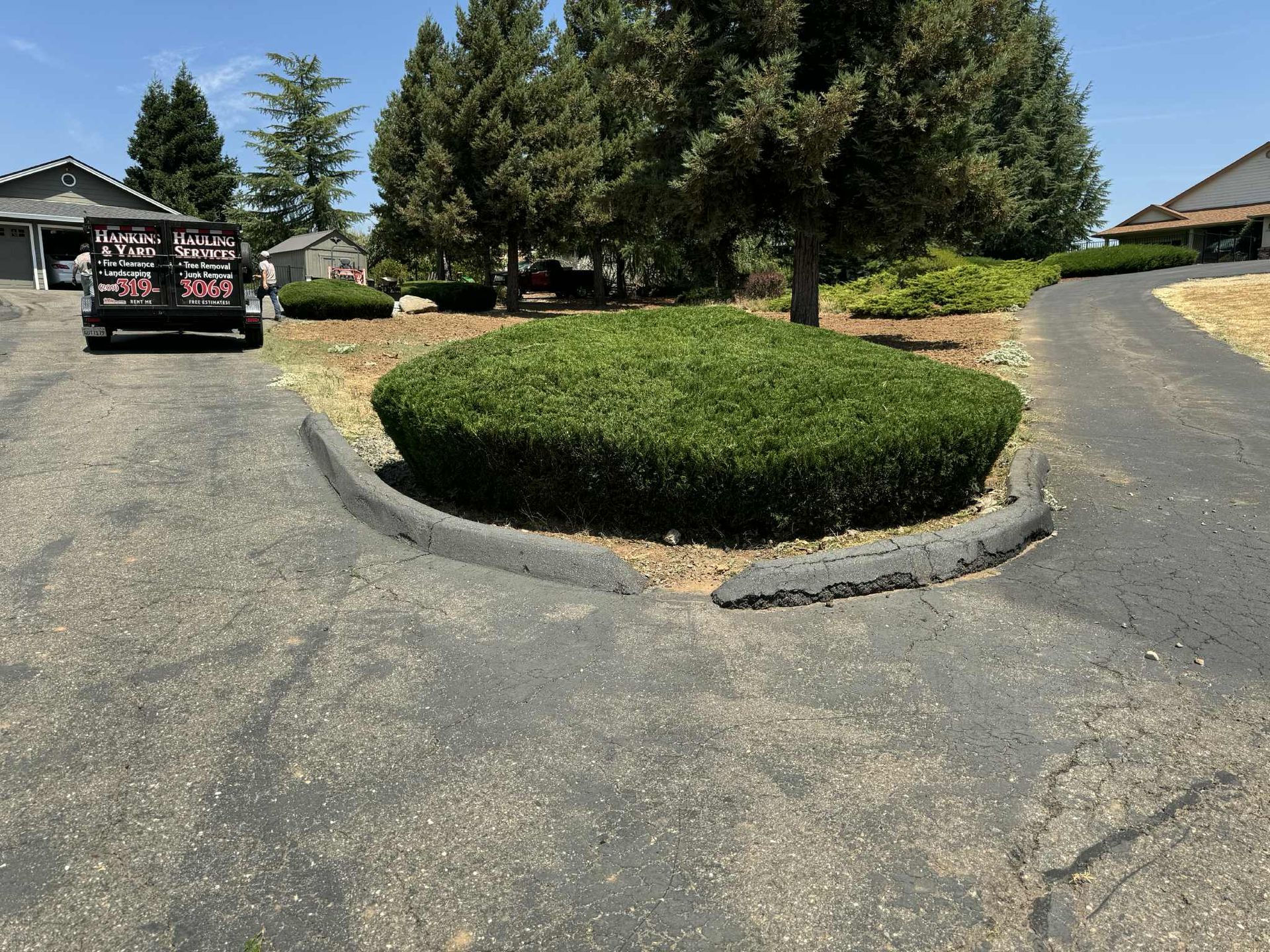 Split driveway with a circular, green bush in the center; a trailer is parked nearby.