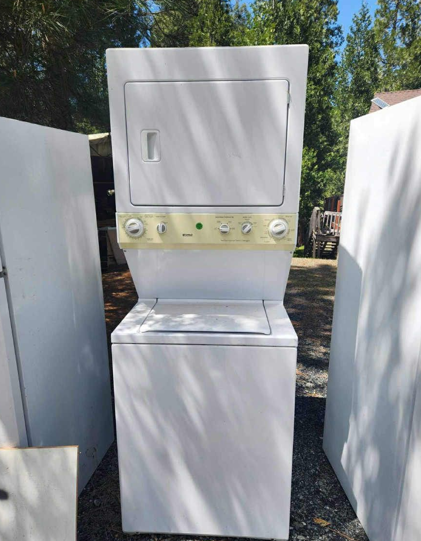 Stacked white washer and dryer with cream-colored control panel, set outside near a forest.