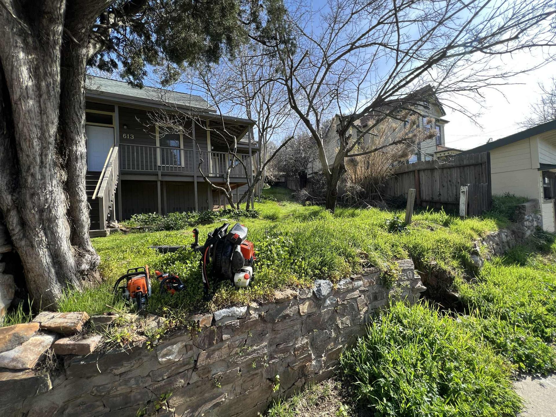 Lawn with overgrown grass, a building, and a chainsaw on a retaining wall on a sunny day.