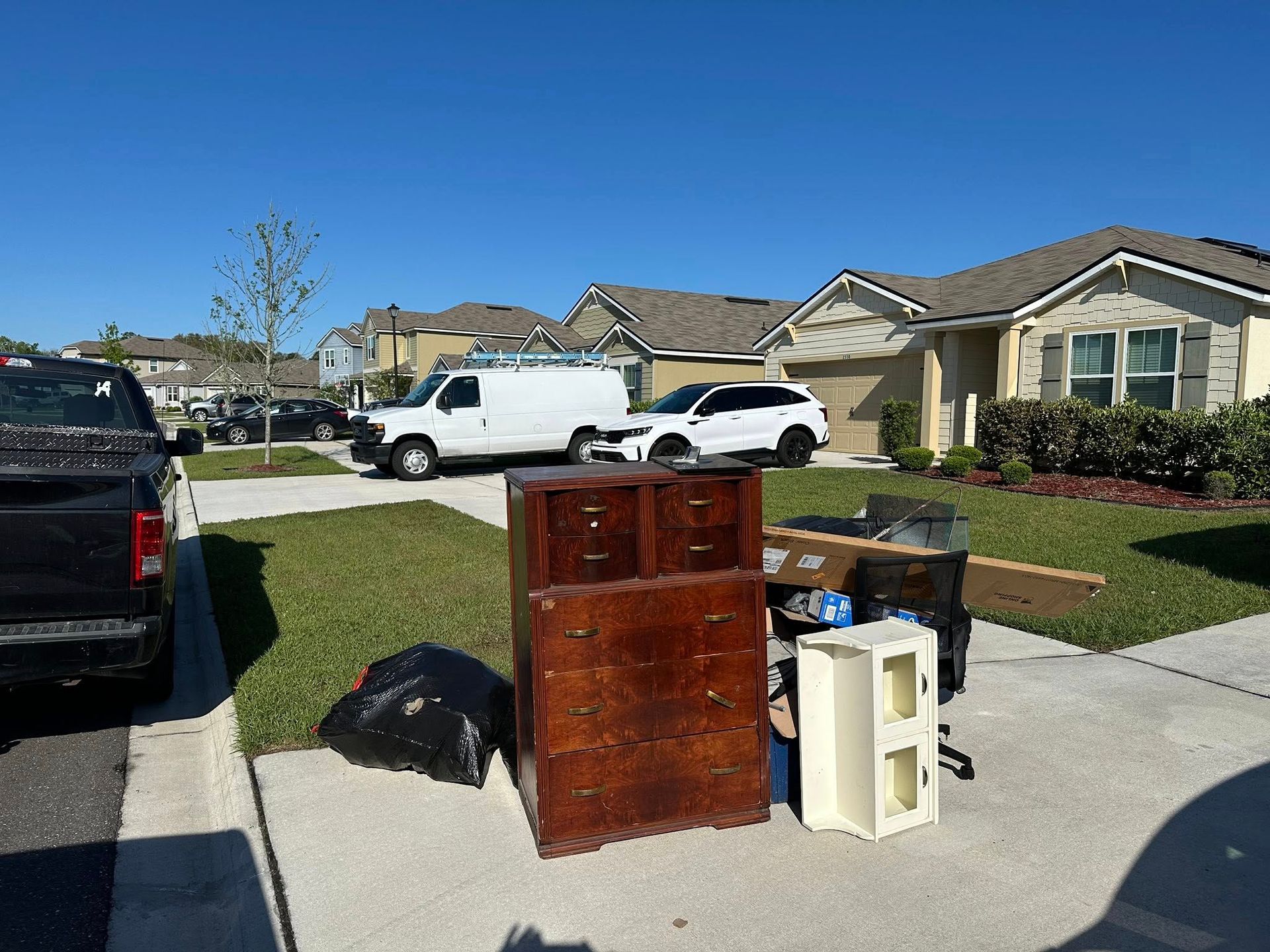 Furniture and trash piled on a driveway in front of suburban houses on a sunny day.