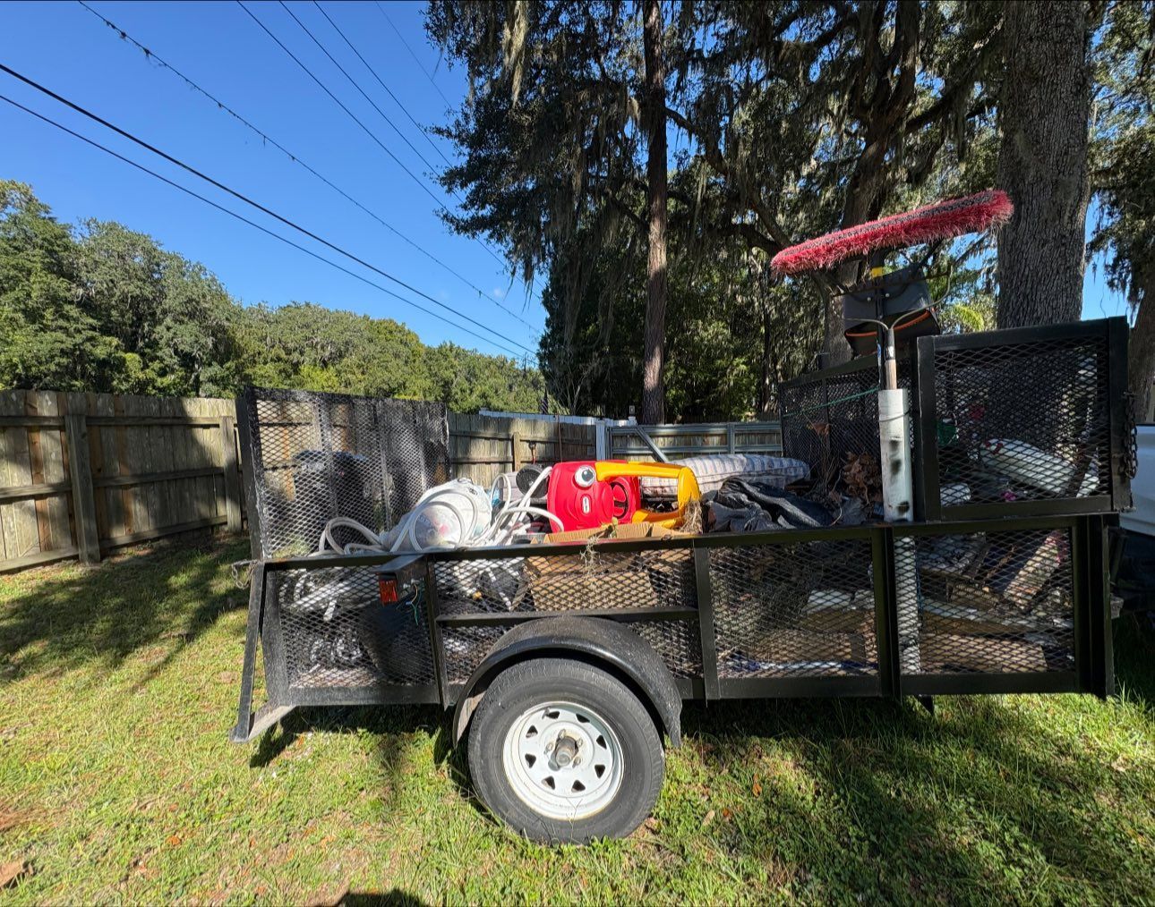 A black trailer filled with various items sits on grass. A brush sticks out.
