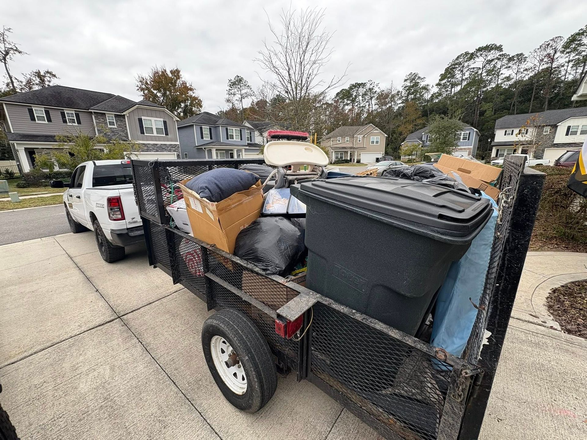 A truck towing a trailer overflowing with various items, including a trash can, on a driveway in a residential area.