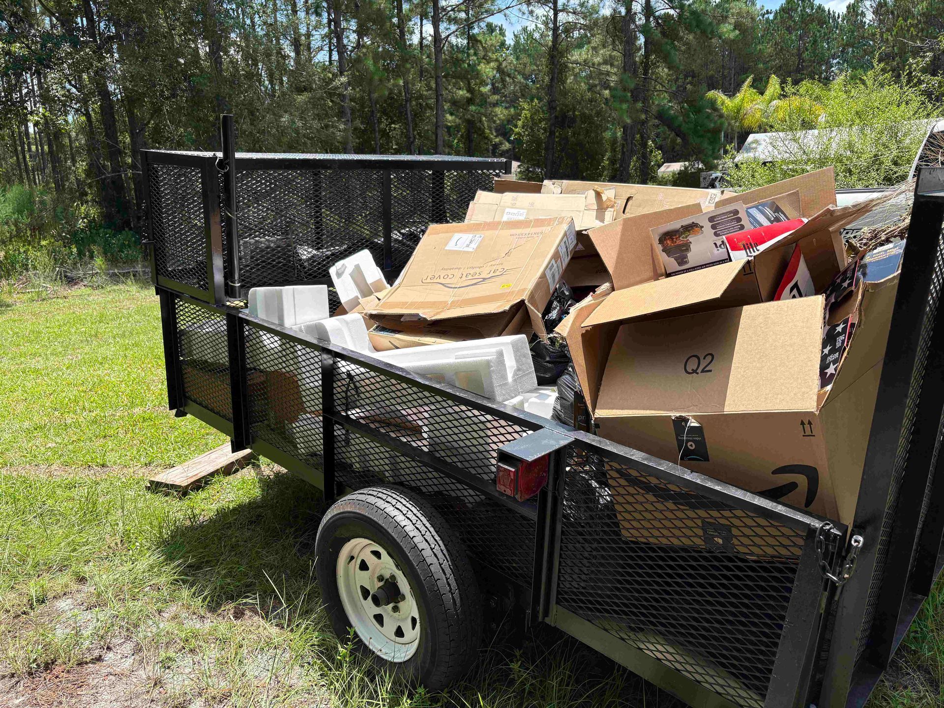Trailer filled with cardboard boxes, trash, and an old computer on grass, under the sun.