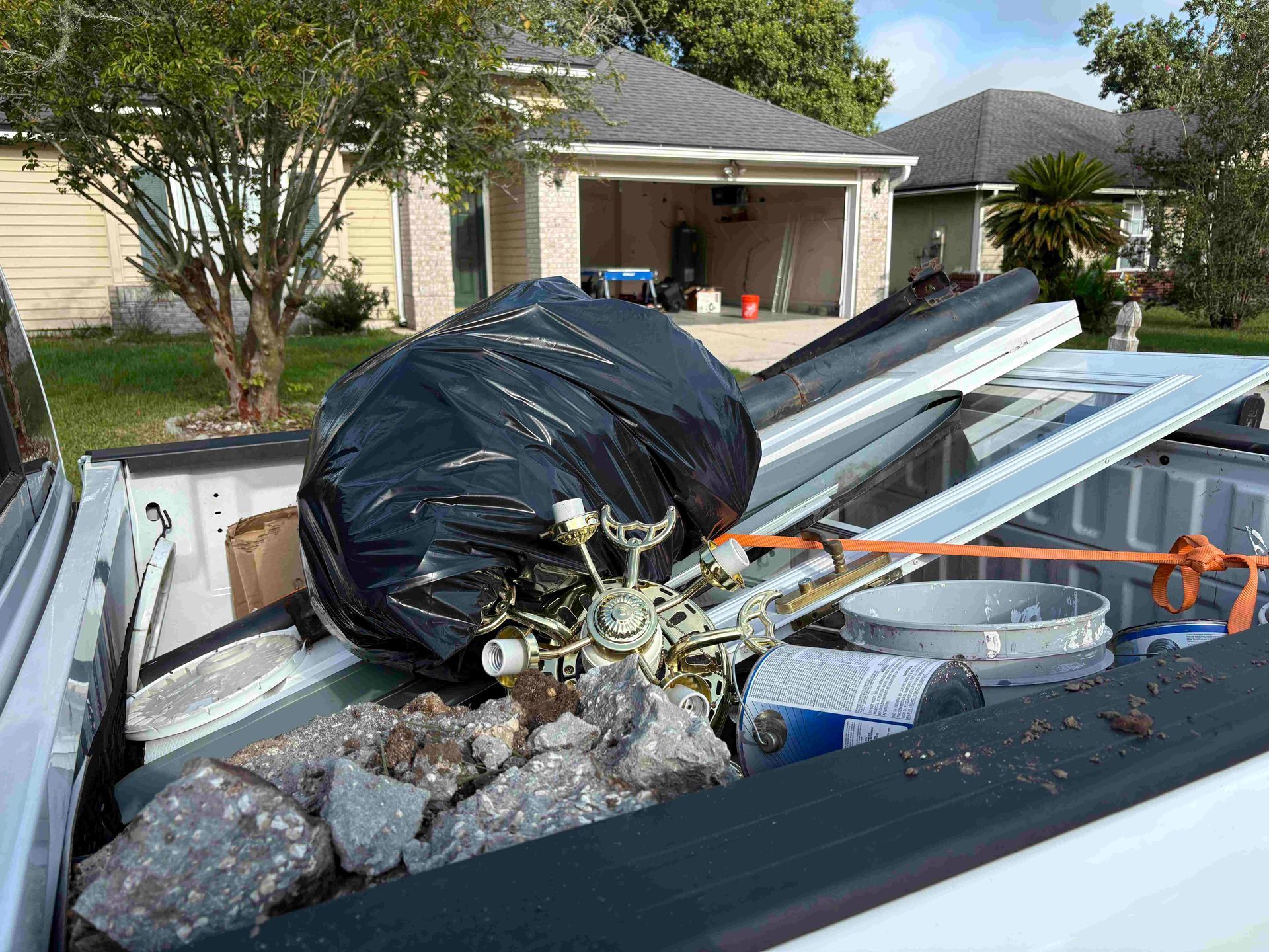 Truck bed filled with trash: black bag, old ceiling fan, construction debris, and a garage in the background.