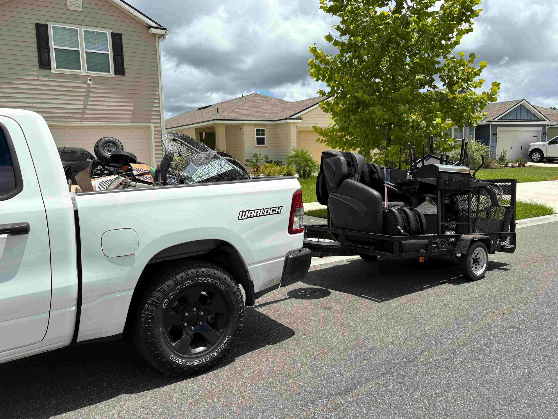 White pickup truck towing a black trailer loaded with equipment on a sunny residential street.