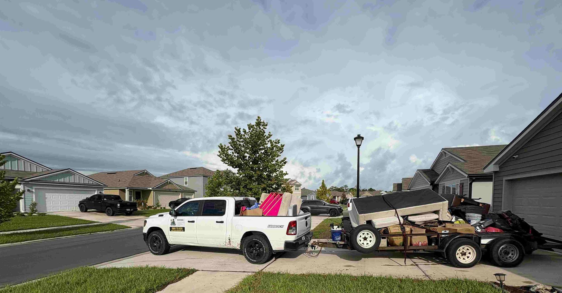White pickup truck with a trailer loaded with furniture parked in front of a house on a cloudy day.