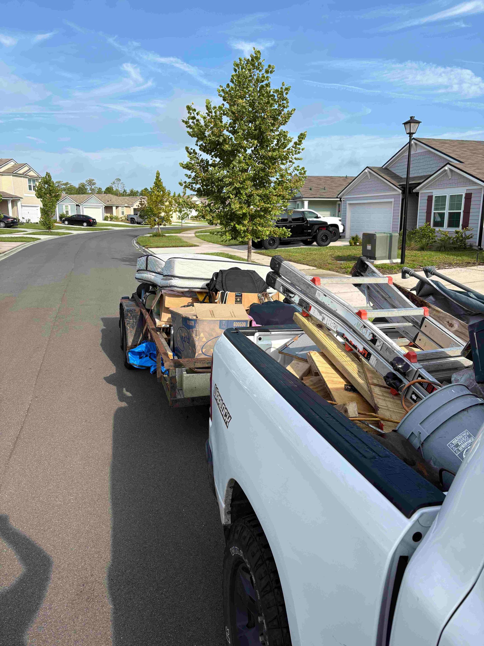 White pickup truck towing a trailer loaded with construction materials on a suburban street.