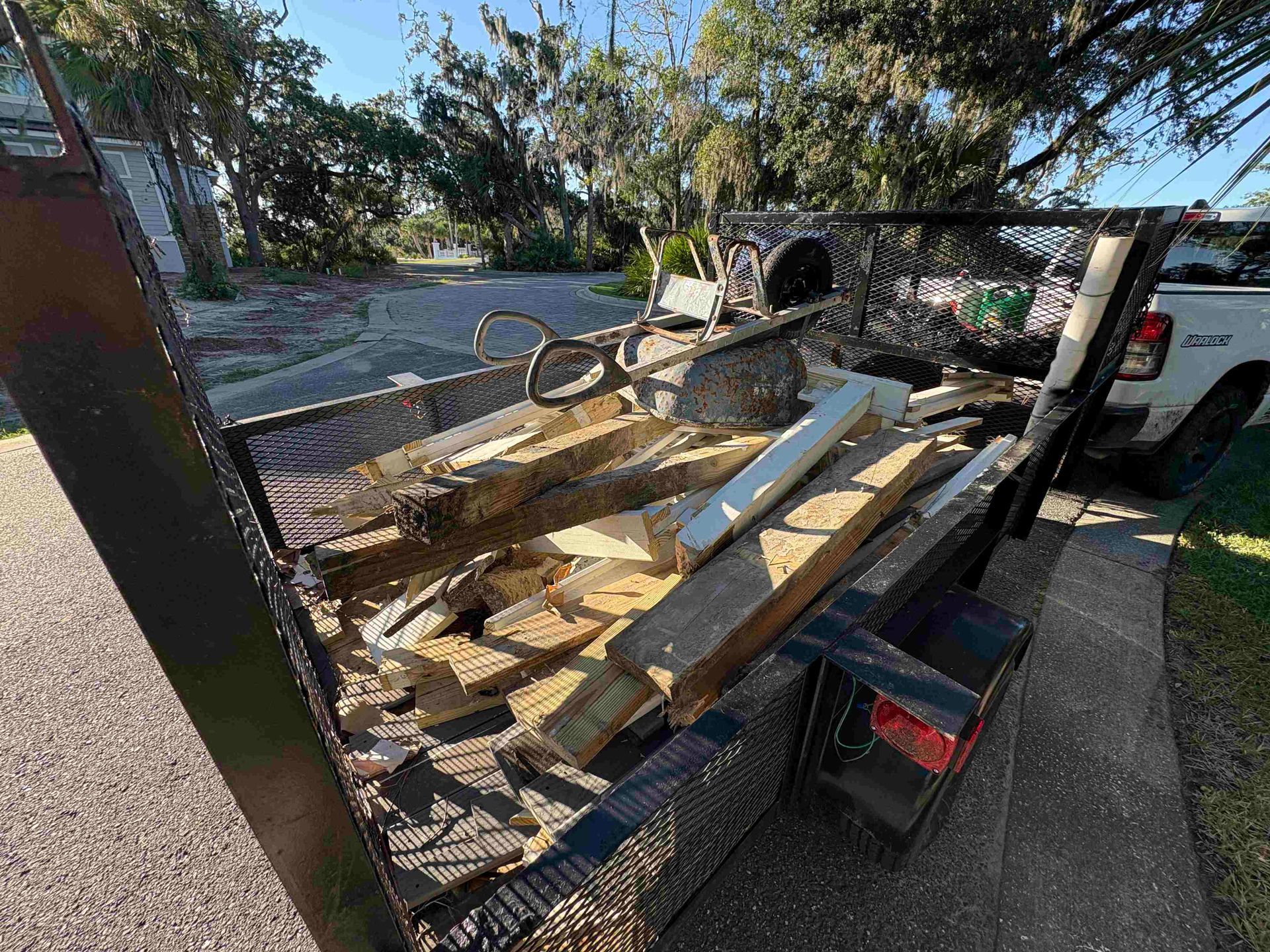 A trailer filled with wooden planks and debris parked on a driveway next to a white pickup truck.