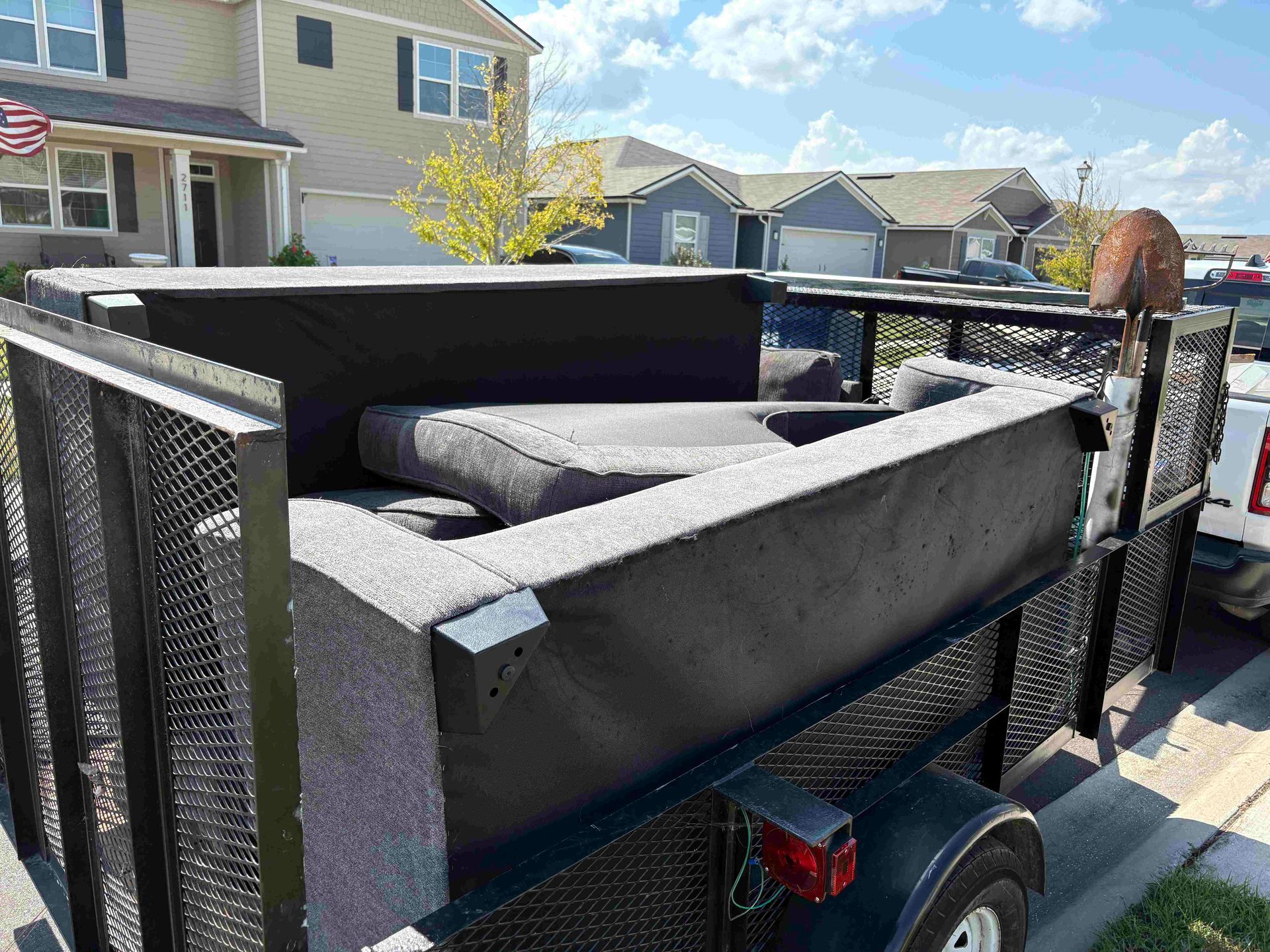 A grey sofa being transported in a black trailer on a sunny day in a suburban neighborhood.