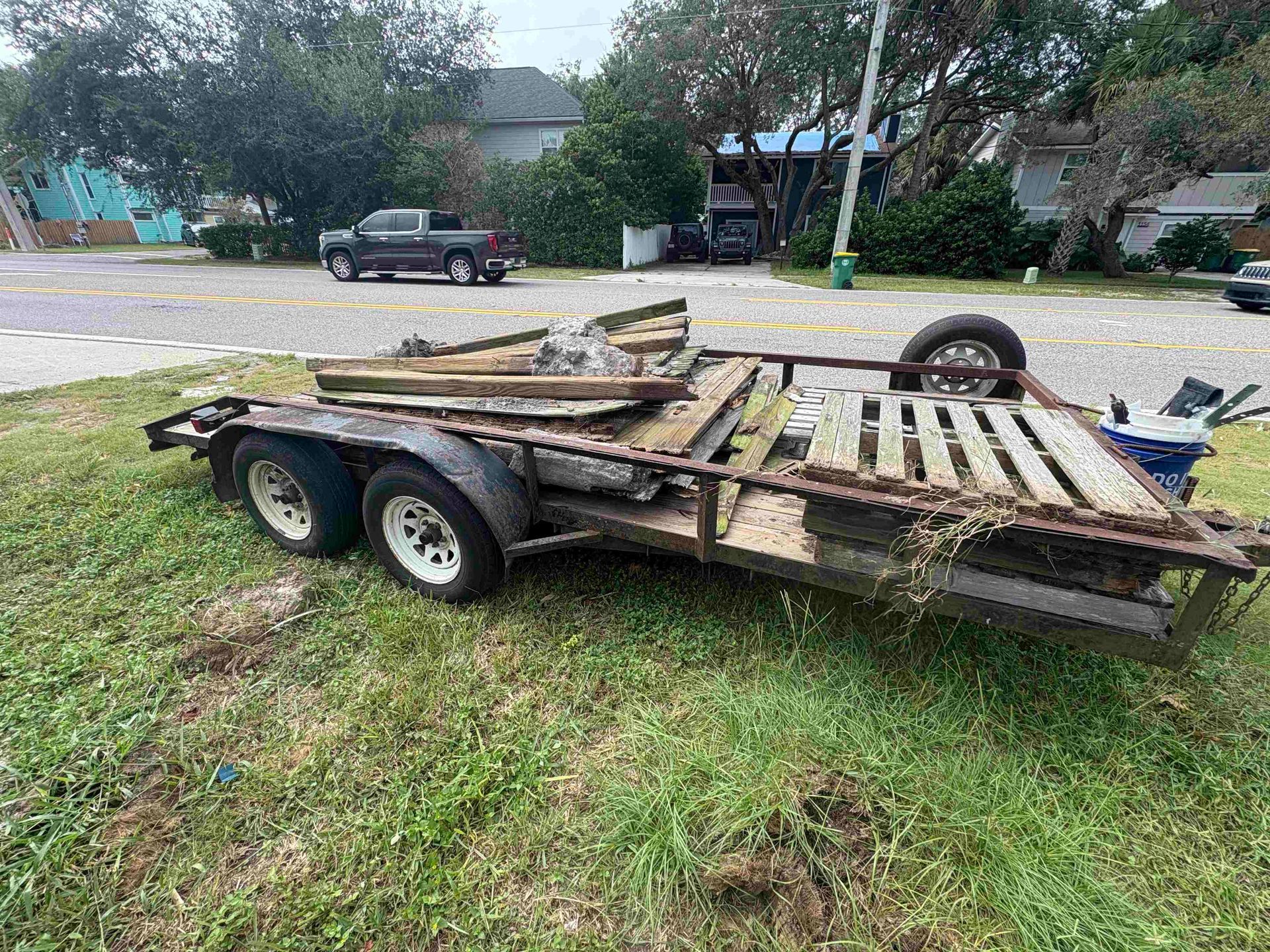 A trailer with debris on a grassy area next to a road. Trees and houses are in the background.