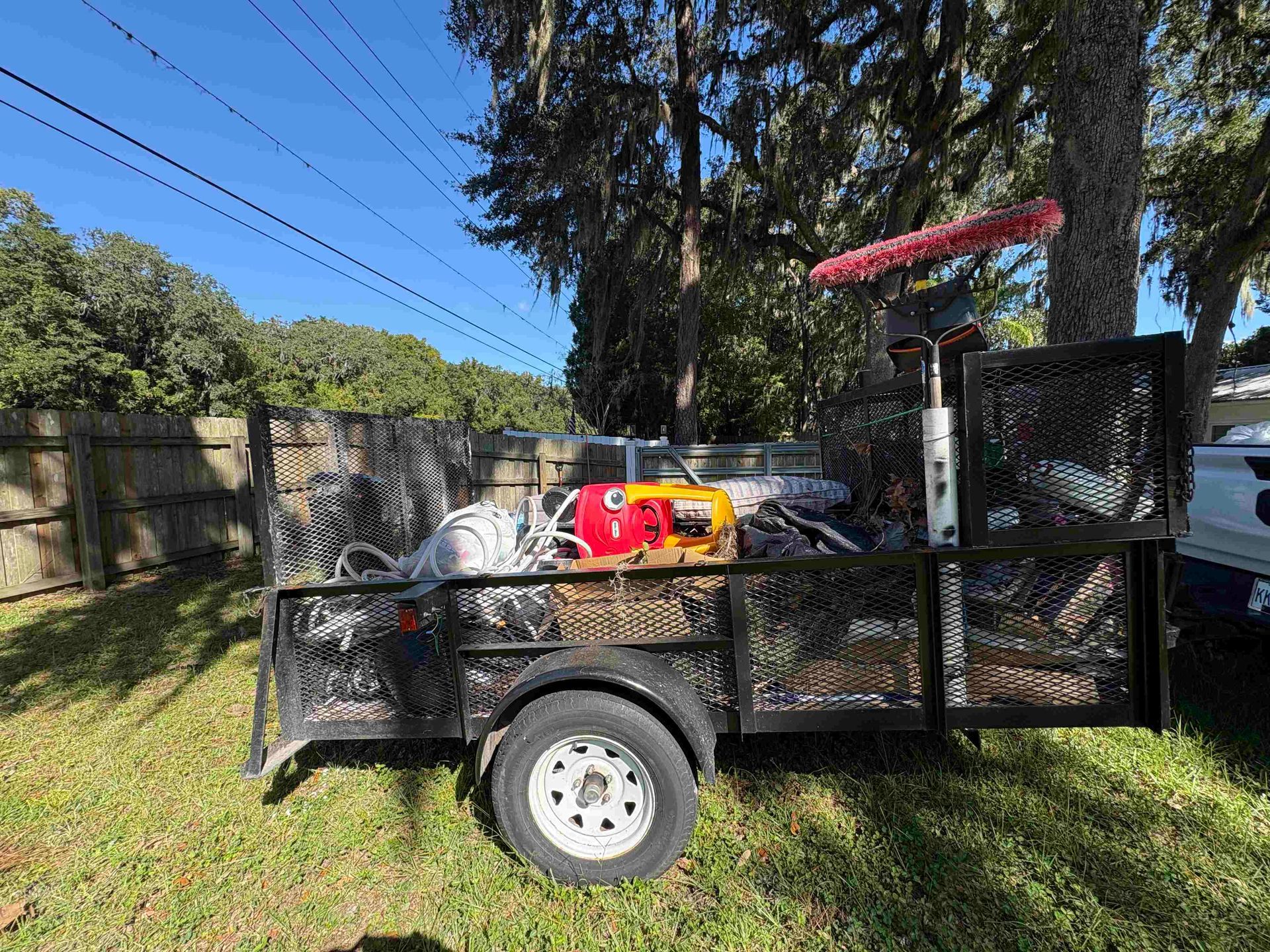 Black utility trailer loaded with equipment, parked on grass next to a wooden fence and trees.