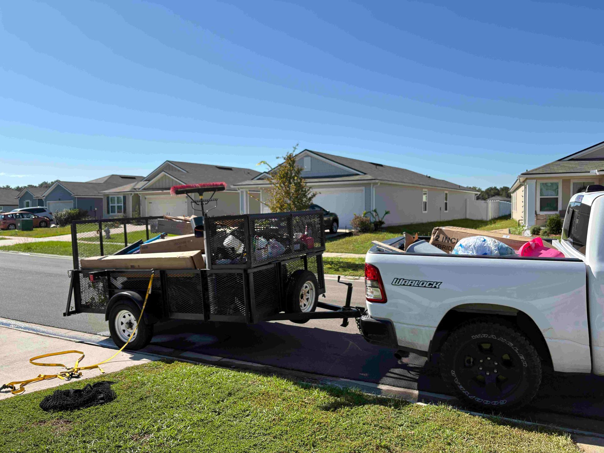 White truck towing a trailer loaded with various items on a residential street under a blue sky.