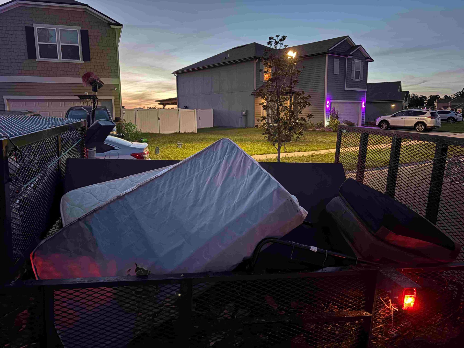Trailer loaded with mattresses and furniture in front of houses at dusk.