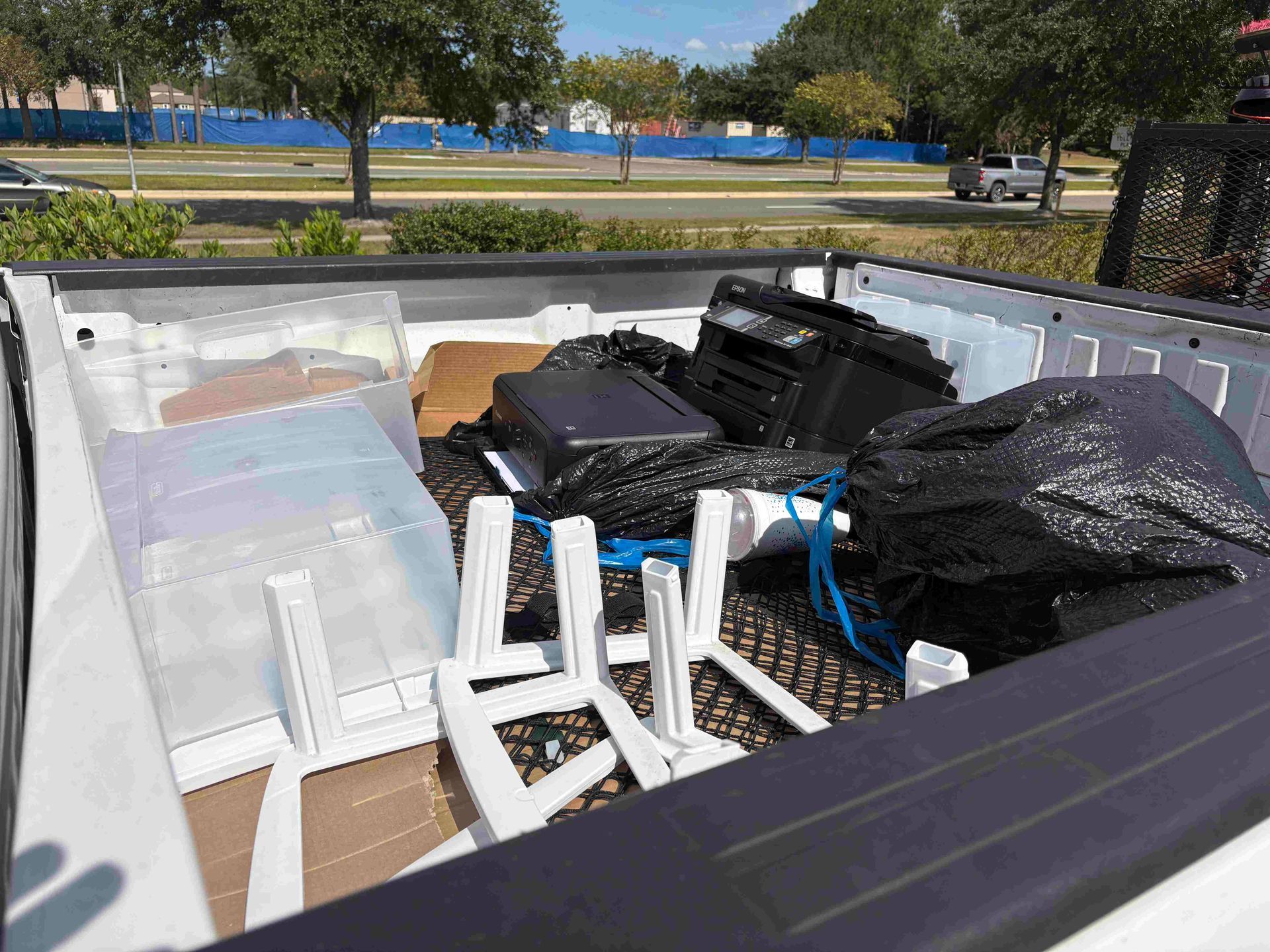 White pickup truck bed filled with assorted trash, including white chairs, bags, and electronics on a sunny day.