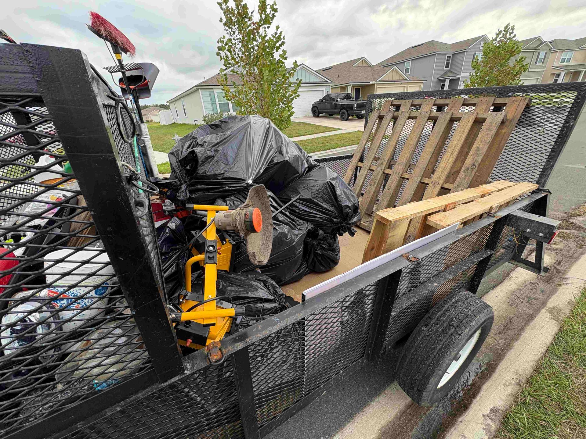 Black trailer filled with trash bags, wood pallets, and tools parked on a residential street.