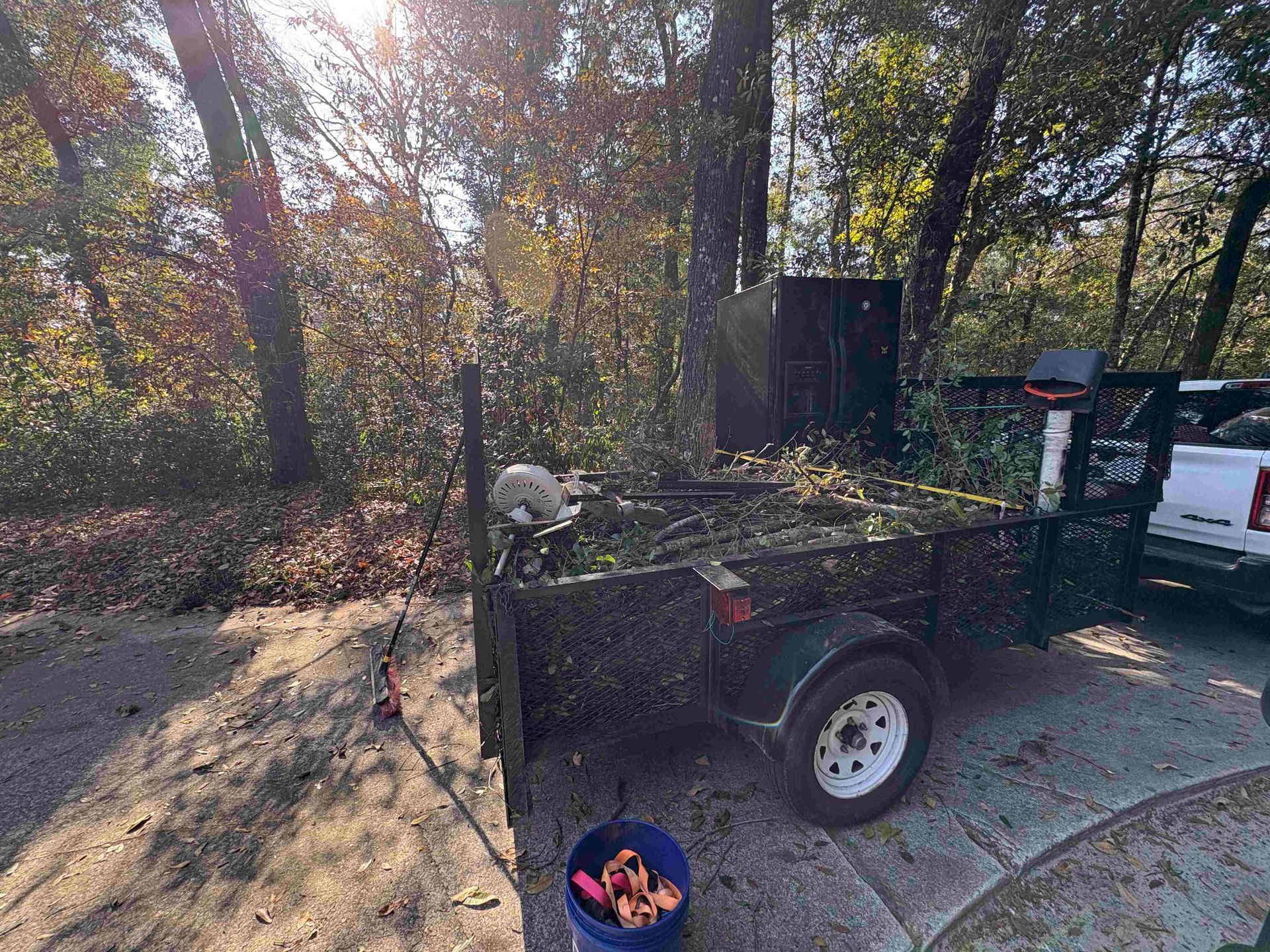 A trailer filled with branches sits on a driveway with a bucket of pink items in front of it.