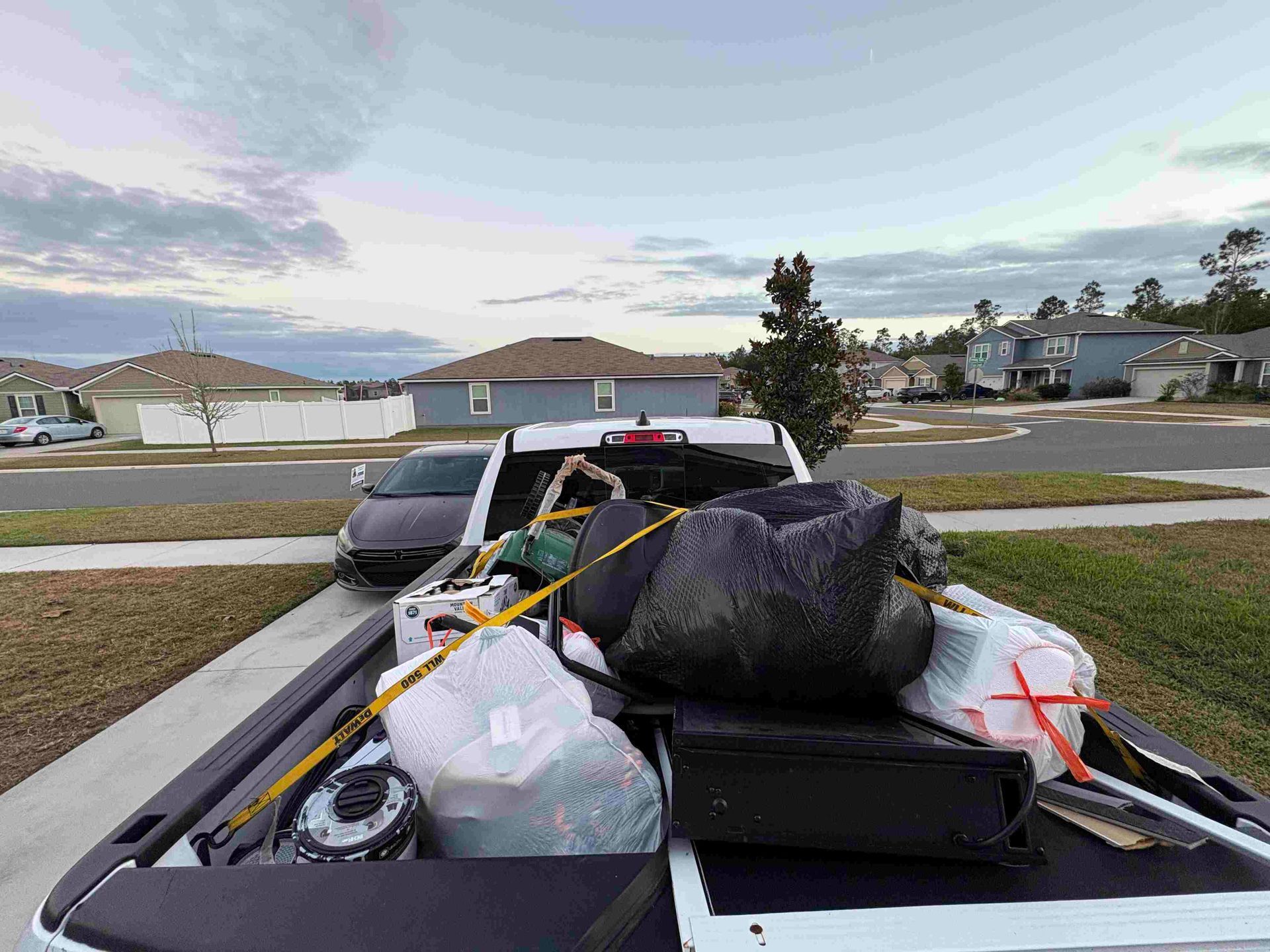 Truck bed filled with trash bags and items, parked in a suburban driveway under a cloudy sky.
