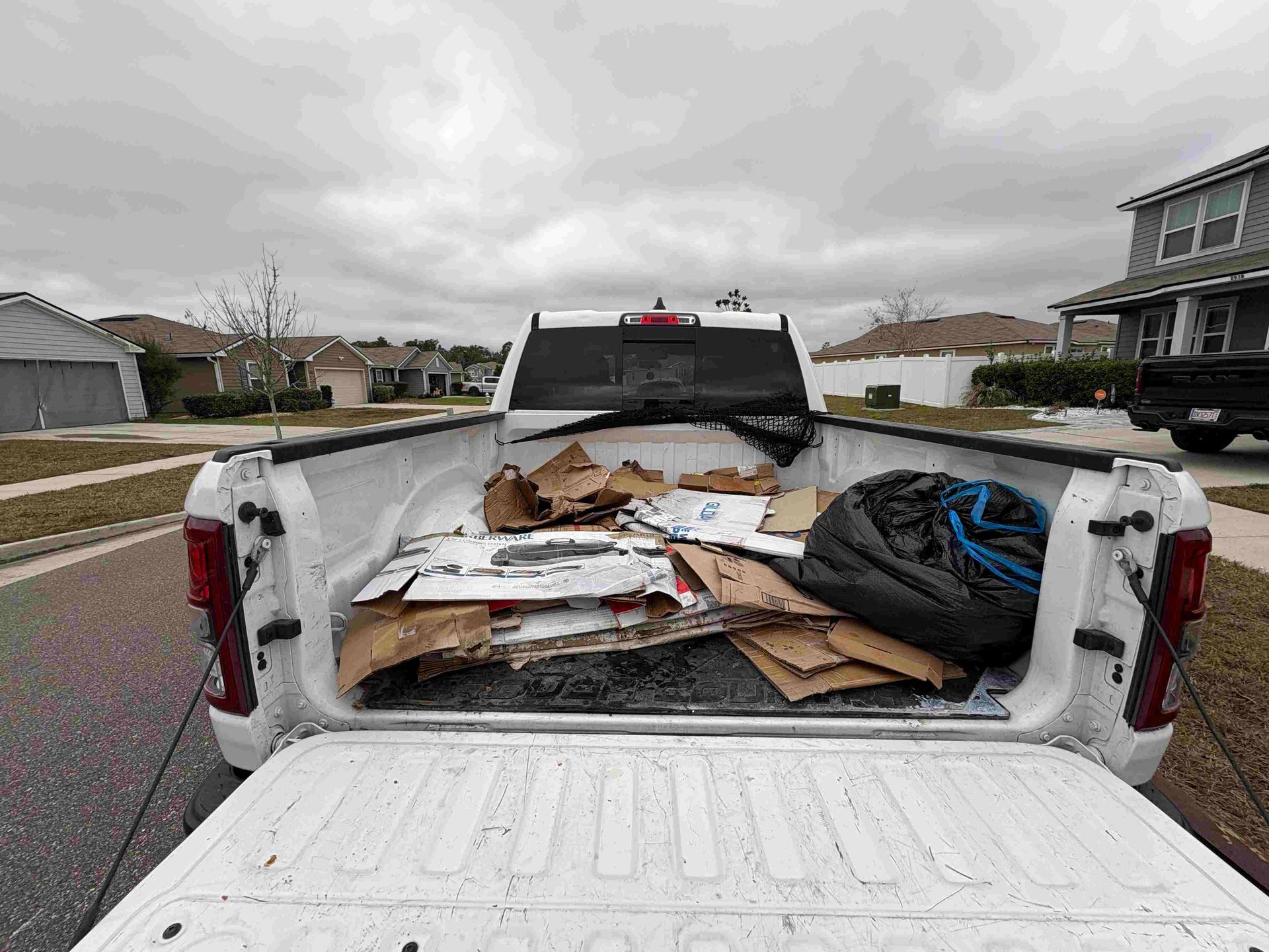 White pickup truck bed filled with cardboard and a black trash bag, parked on a street in a residential area.