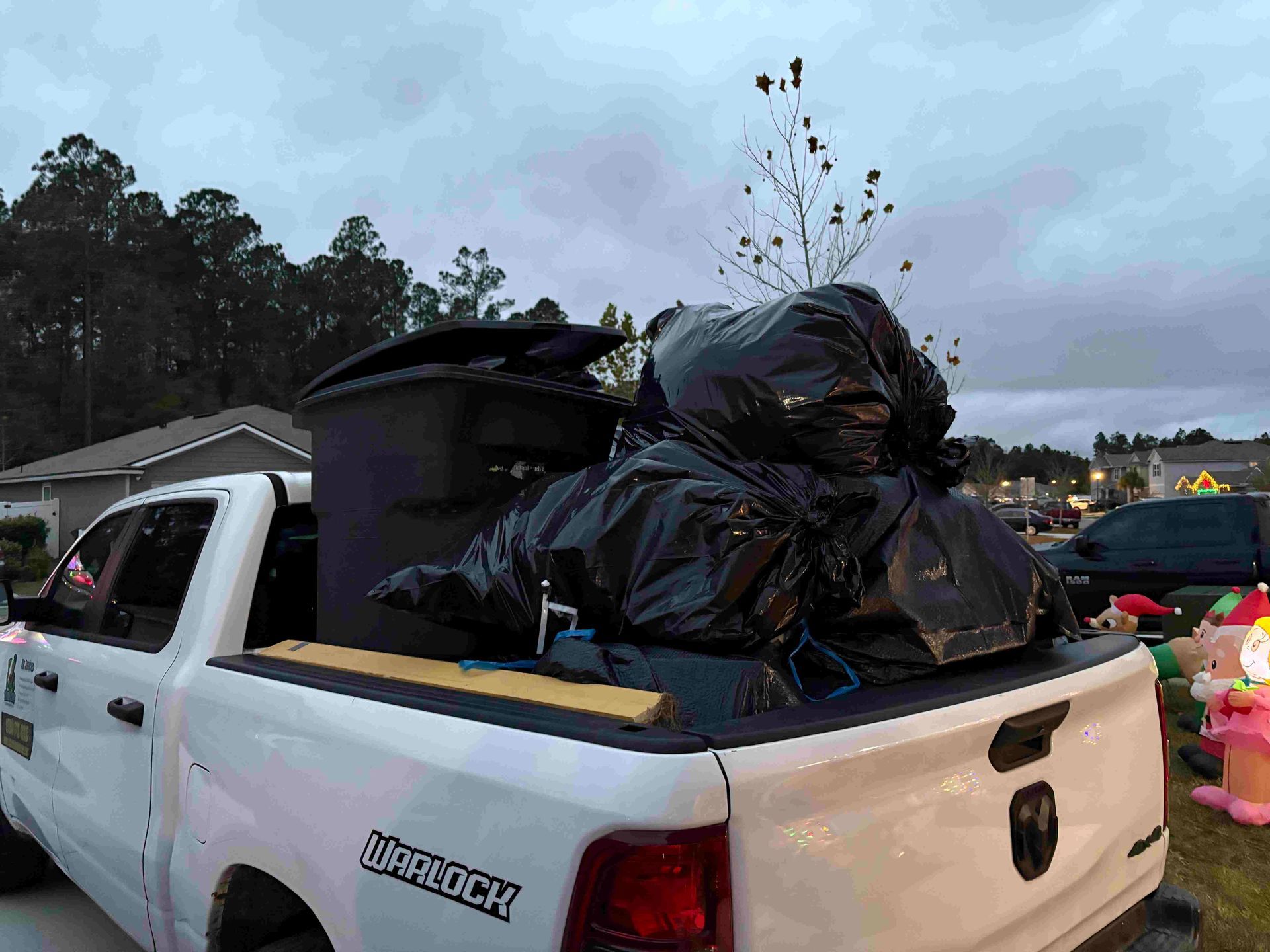 White pickup truck bed filled with black trash bags and a trash can, set against a residential backdrop.