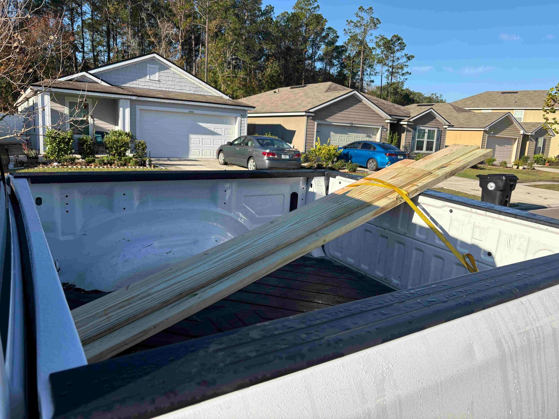 Pickup truck bed holding long wooden planks, secured with a yellow strap. Houses in the background.