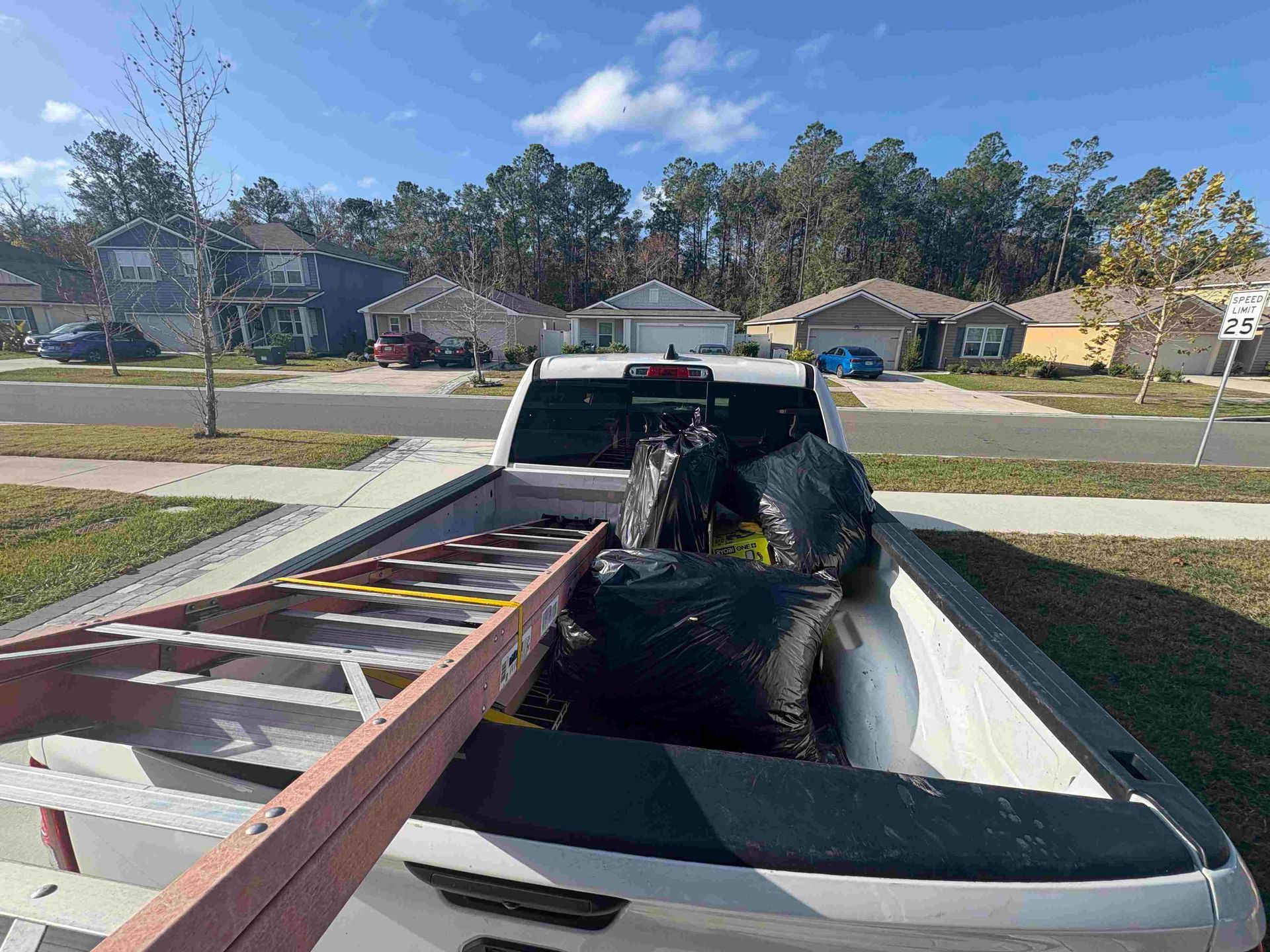 Pickup truck bed loaded with black bags and a ladder, parked on a residential street.
