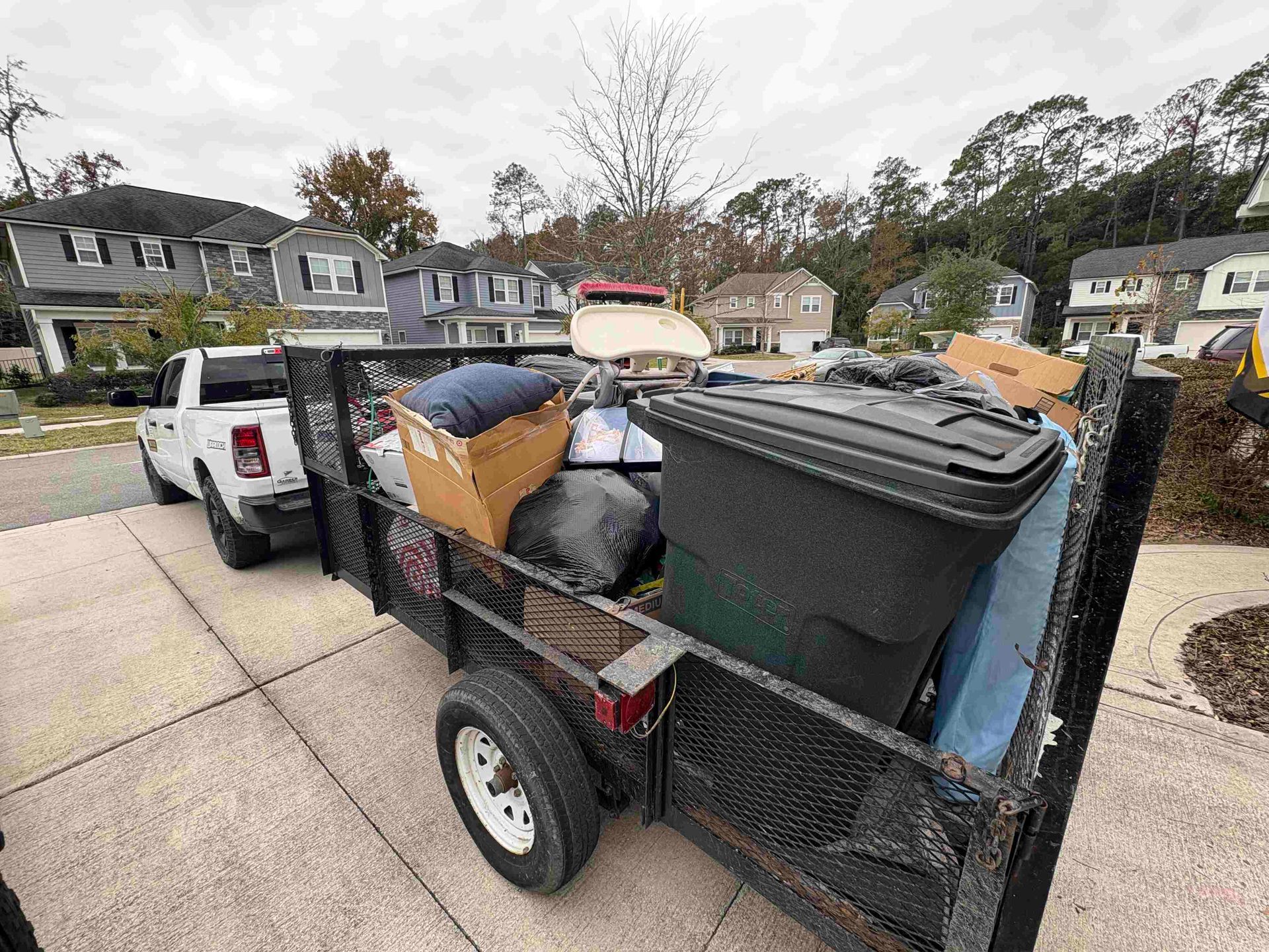 White pickup truck pulling a trailer loaded with household items on a residential street.