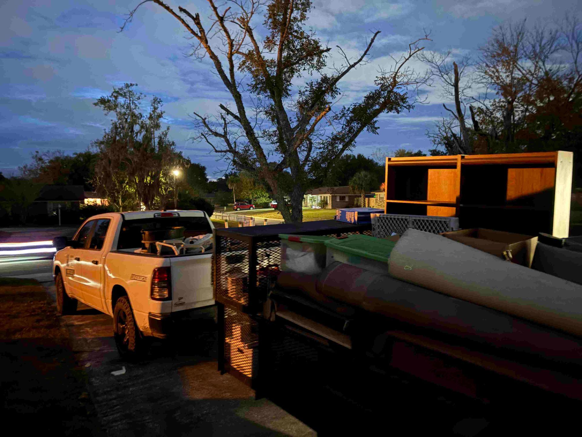 White truck towing a trailer with rolled items; parked on a street in a residential area at dusk.