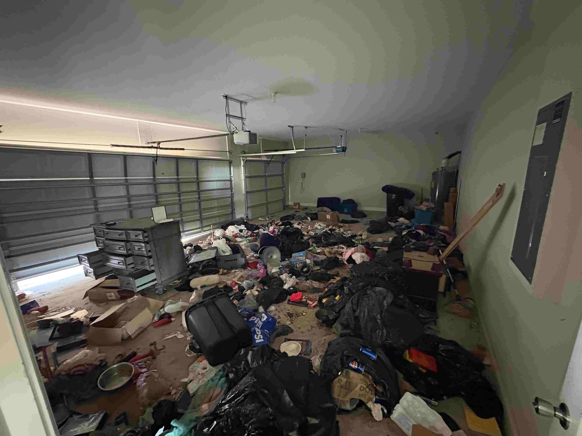Messy garage with debris and trash bags scattered on the floor. Garage door open.