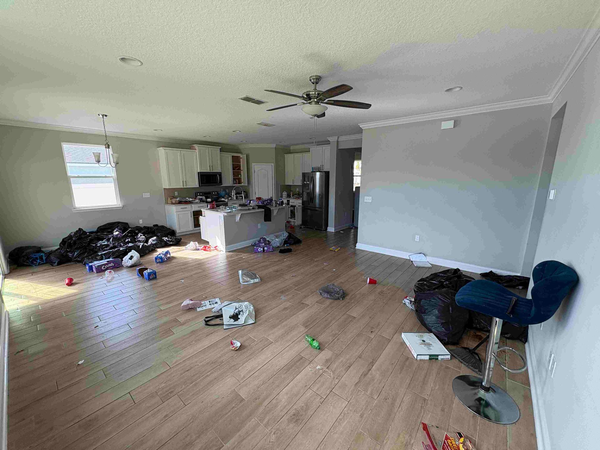Cluttered interior with debris on floor, kitchen in background. Ceiling fan and bar stool visible.