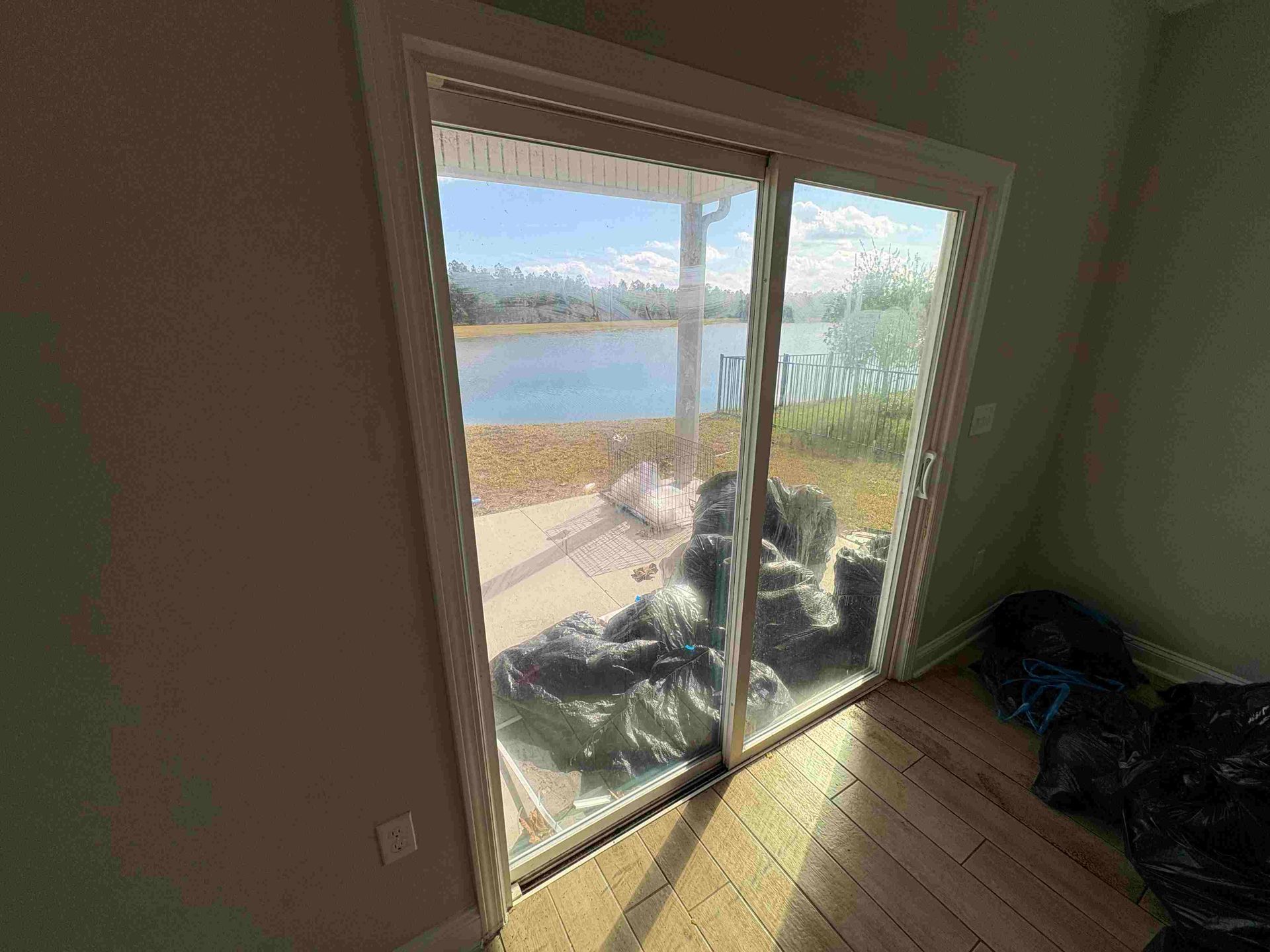 Sliding glass door with a view of a lake and a patio. Black bags are inside. Sunlit interior.