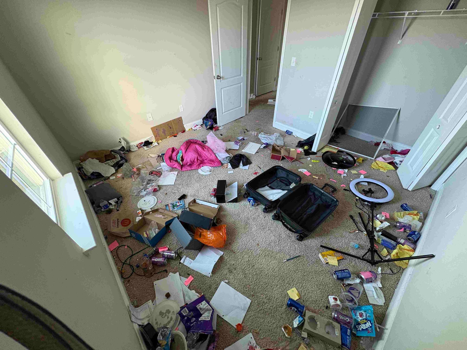 A messy room: clothes, luggage, and trash scattered across the floor, with a partially open closet and door.