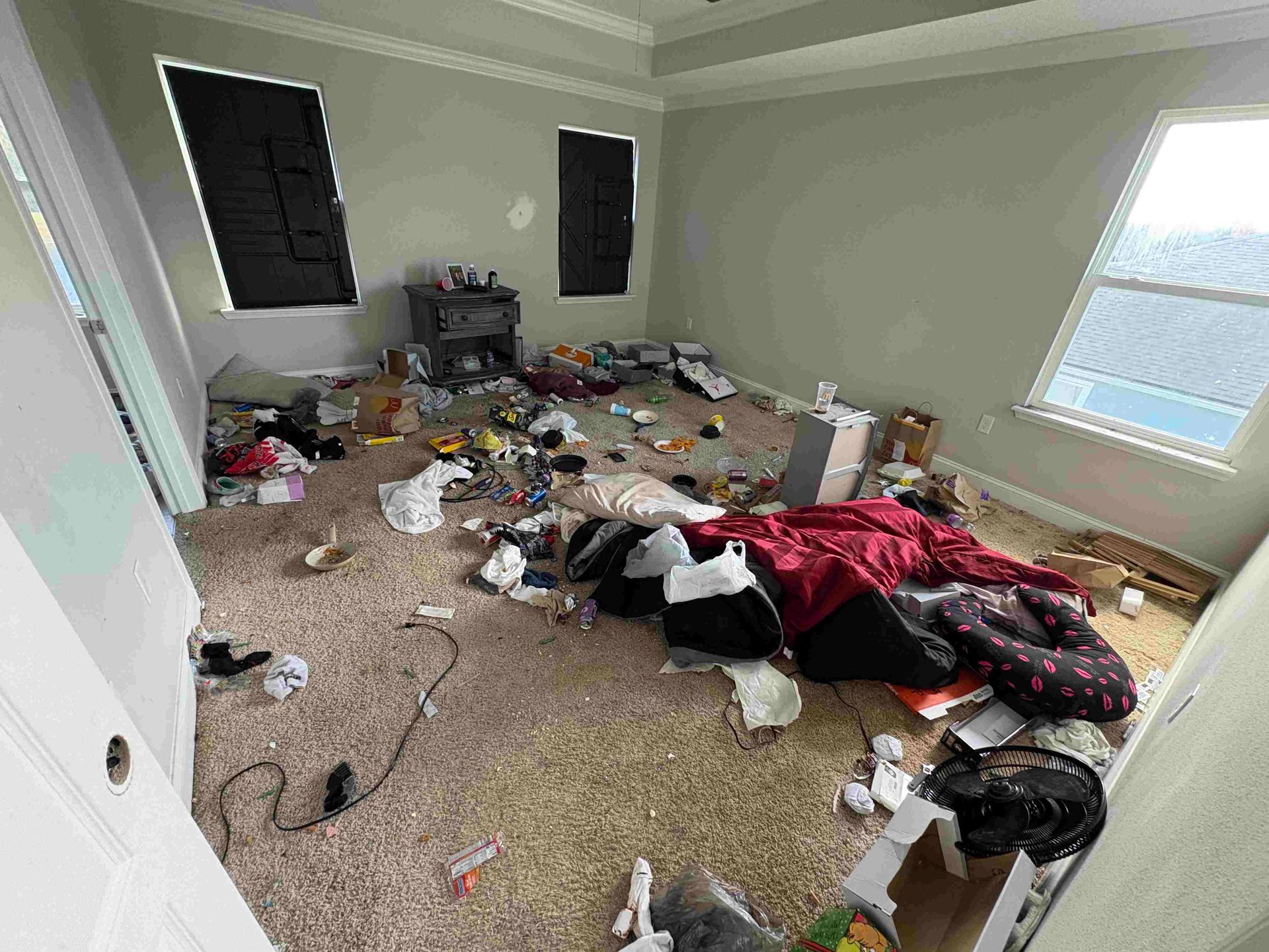 Messy bedroom with trash and items scattered on the floor; two small windows covered in black, one large window open.