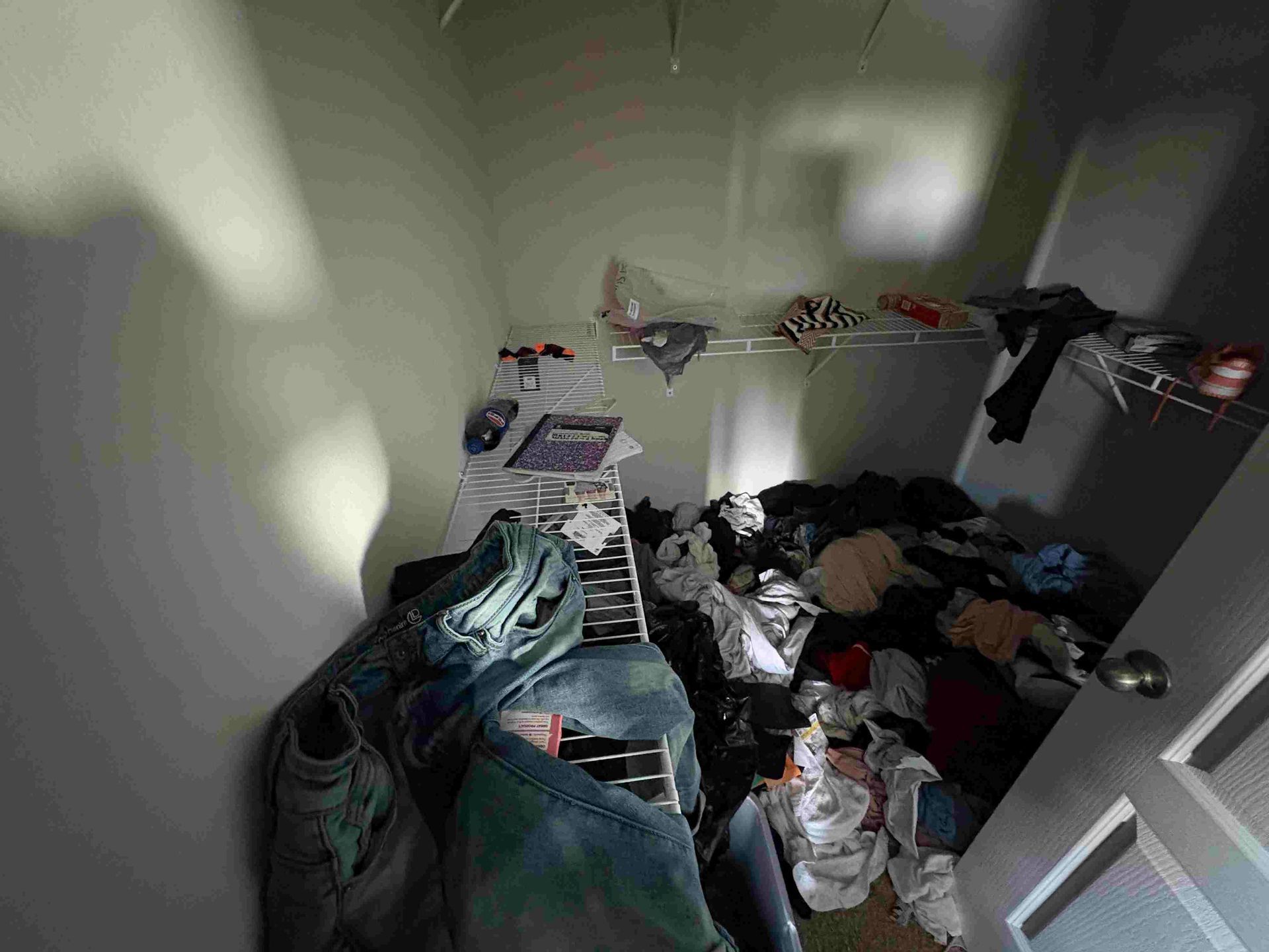 Messy closet with clothes piled on the floor and shelves; door on the right.