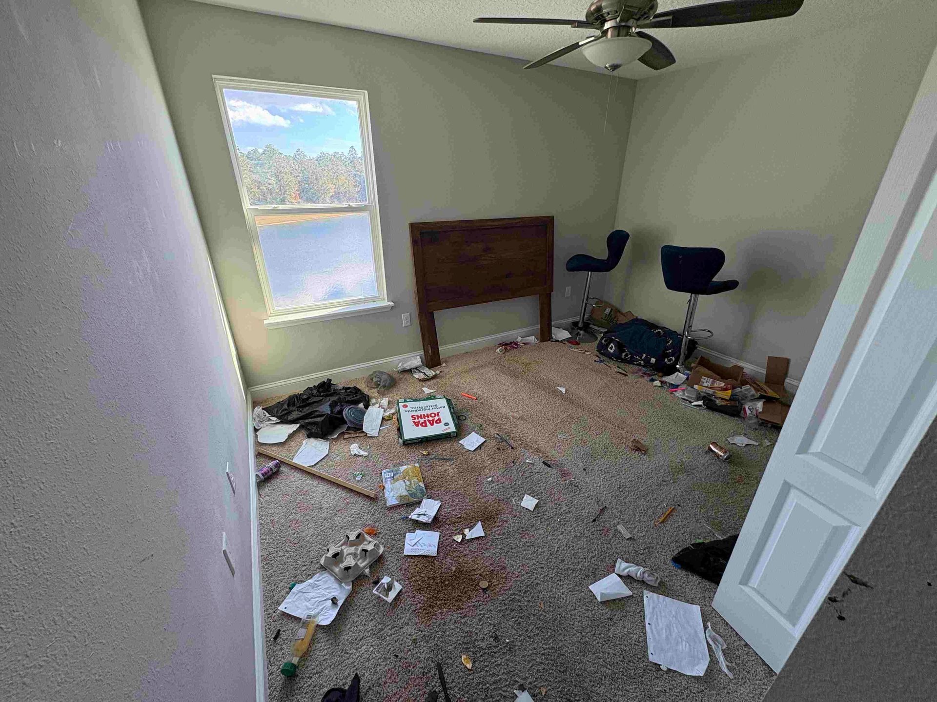 Messy bedroom with trash on the floor, headboard, window, two chairs.