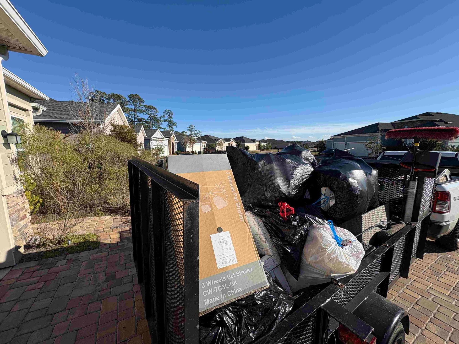 Trailer filled with trash bags, boxes, and items, parked on a driveway in a sunny residential neighborhood.