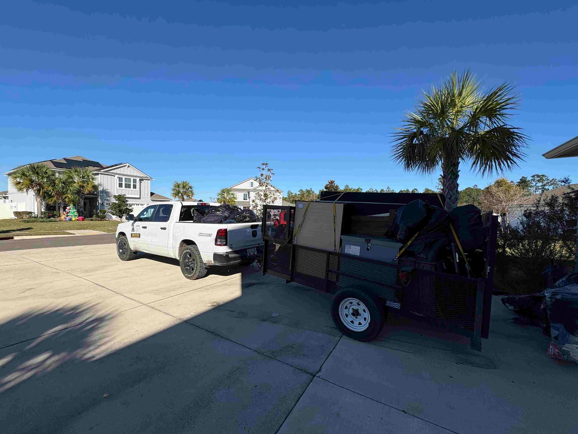 White truck towing a dark trailer in a driveway under a blue sky, near palm trees and houses.