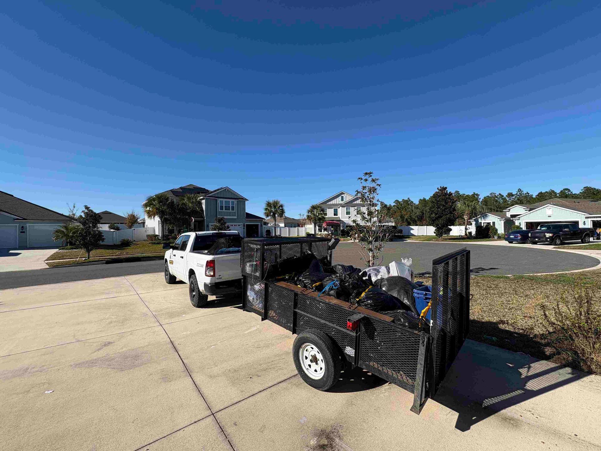 White truck towing a black trailer filled with yard waste, parked on a driveway in a sunny suburban neighborhood.