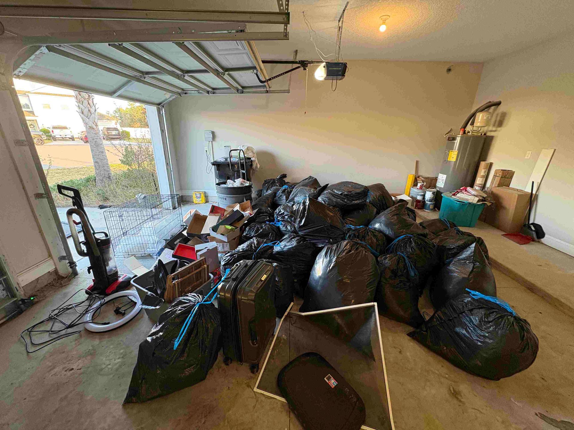 Garage filled with black trash bags, cardboard boxes, and various items. Garage door open.